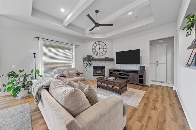 living room featuring a raised ceiling, ceiling fan, beamed ceiling, light hardwood / wood-style floors, and a tiled fireplace