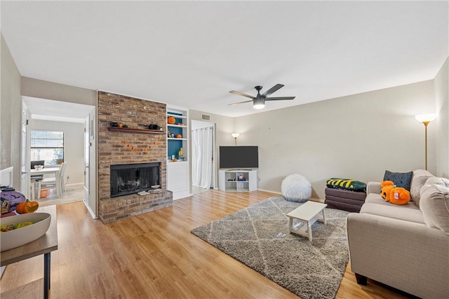 living room featuring ceiling fan, wood-type flooring, built in shelves, and a brick fireplace