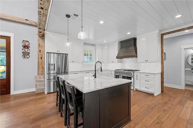 kitchen with appliances with stainless steel finishes, custom exhaust hood, white cabinetry, and a kitchen island with sink