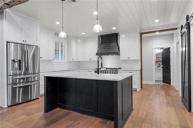 kitchen featuring custom exhaust hood, a barn door, a center island with sink, stainless steel refrigerator with ice dispenser, and light stone counters