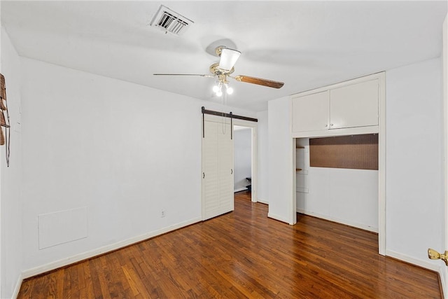 unfurnished bedroom with a barn door, ceiling fan, a closet, and dark wood-type flooring