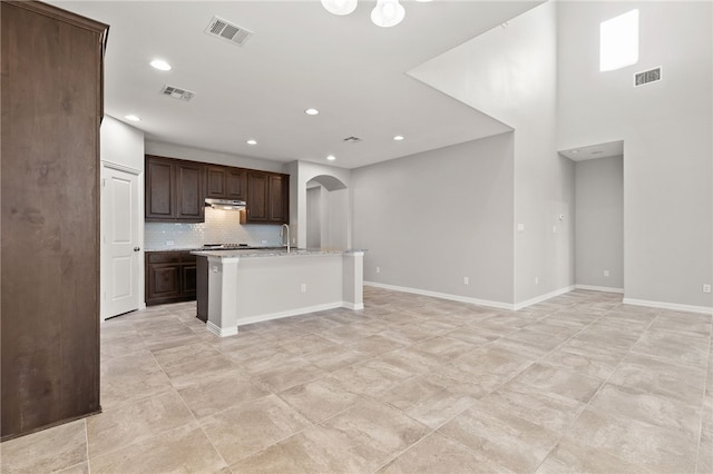 kitchen featuring backsplash, light stone counters, dark brown cabinetry, an island with sink, and light tile patterned flooring