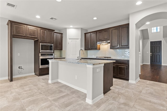 kitchen with dark brown cabinetry, sink, light stone counters, a kitchen island with sink, and appliances with stainless steel finishes