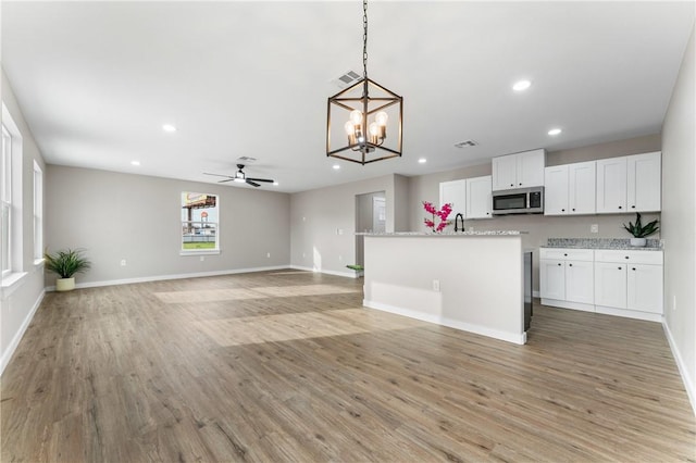 kitchen with light wood finished floors, stainless steel microwave, ceiling fan with notable chandelier, hanging light fixtures, and white cabinetry