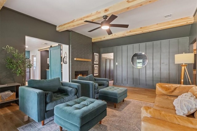 living room with a brick fireplace, ceiling fan, wood-type flooring, a barn door, and lofted ceiling with beams