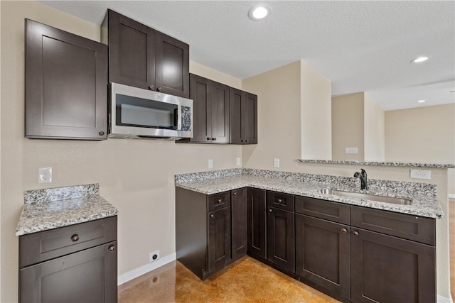 kitchen with stainless steel microwave, a peninsula, light stone countertops, dark brown cabinets, and a sink