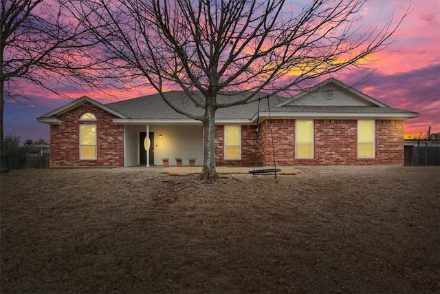 back of house at dusk featuring roof with shingles, fence, and brick siding