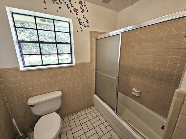 bathroom with tile patterned floors, bath / shower combo with glass door, a textured ceiling, tile walls, and toilet