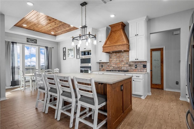 kitchen featuring premium range hood, pendant lighting, a tray ceiling, a center island with sink, and white cabinets