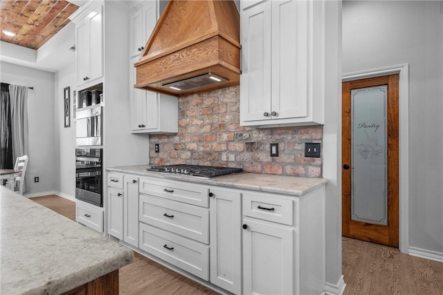 kitchen with stainless steel gas stovetop, custom exhaust hood, light hardwood / wood-style flooring, white cabinetry, and brick ceiling