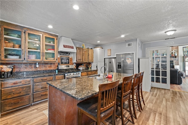 kitchen featuring light wood-type flooring, a textured ceiling, a kitchen island with sink, appliances with stainless steel finishes, and custom exhaust hood