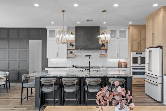 kitchen with custom exhaust hood, an island with sink, a notable chandelier, and white appliances