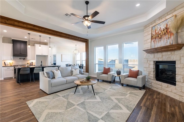 living room with a stone fireplace, ceiling fan with notable chandelier, wood-type flooring, and a tray ceiling