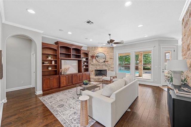 living room featuring dark wood-type flooring, ceiling fan, ornamental molding, and a stone fireplace