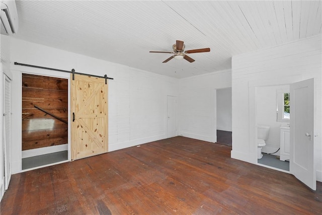 unfurnished bedroom featuring ensuite bath, ceiling fan, dark wood-type flooring, a barn door, and a wall unit AC