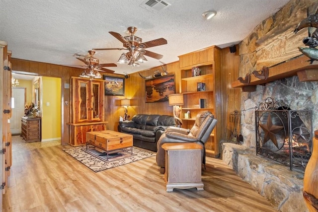 living room with ceiling fan, a stone fireplace, light hardwood / wood-style flooring, a textured ceiling, and wooden walls