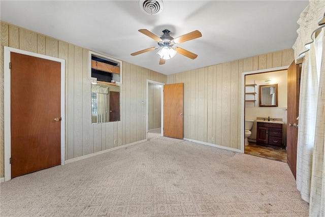 unfurnished bedroom featuring sink, ensuite bathroom, ceiling fan, wood walls, and light colored carpet