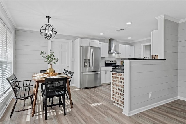 kitchen with wall chimney exhaust hood, white cabinetry, crown molding, decorative light fixtures, and appliances with stainless steel finishes