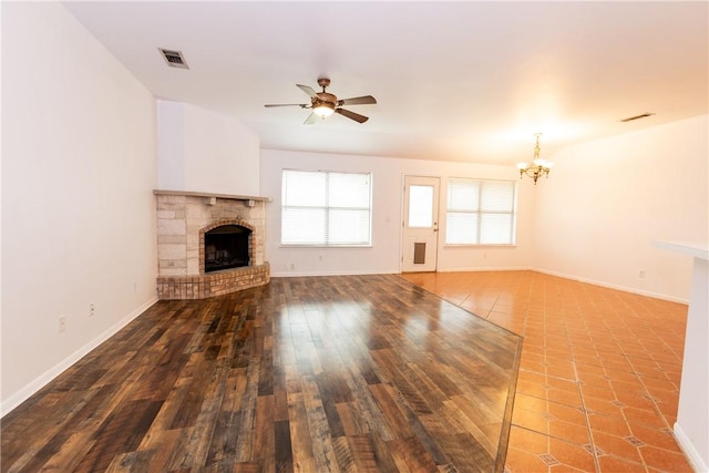 unfurnished living room featuring a brick fireplace, ceiling fan with notable chandelier, visible vents, and baseboards