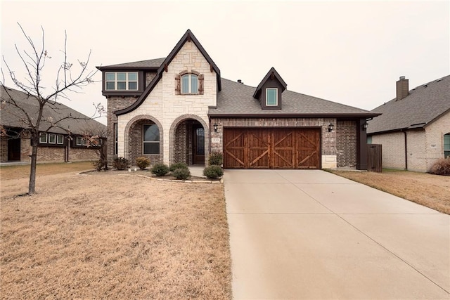 french country inspired facade featuring driveway, stone siding, roof with shingles, a garage, and brick siding