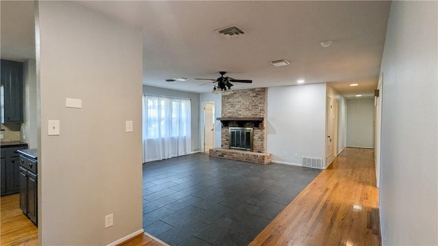 living room featuring dark wood-style floors, visible vents, a brick fireplace, and a ceiling fan