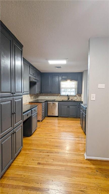 kitchen with under cabinet range hood, a sink, stainless steel dishwasher, light wood finished floors, and dark countertops