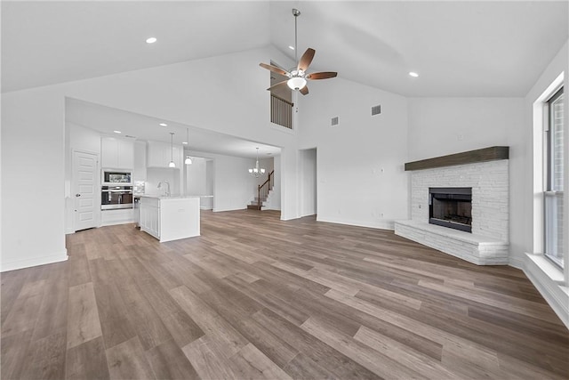 unfurnished living room with hardwood / wood-style floors, high vaulted ceiling, ceiling fan with notable chandelier, a stone fireplace, and sink