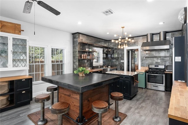 kitchen featuring ceiling fan with notable chandelier, light hardwood / wood-style flooring, wall chimney exhaust hood, appliances with stainless steel finishes, and a kitchen island