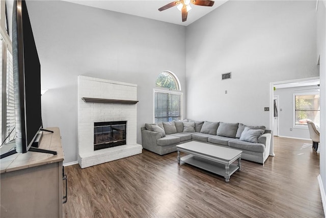 living room with ceiling fan, a fireplace, a towering ceiling, and dark wood-type flooring