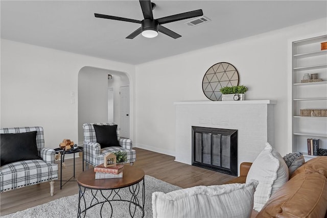 living room featuring built in features, hardwood / wood-style floors, a brick fireplace, and ceiling fan