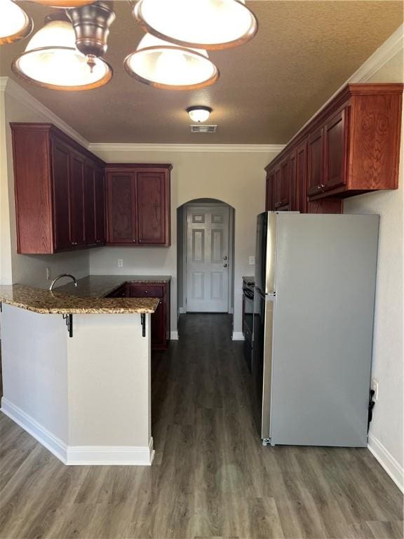 kitchen with dark wood-type flooring, light stone countertops, ornamental molding, kitchen peninsula, and stainless steel refrigerator