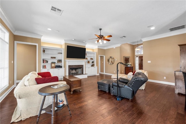 living room with a fireplace, ceiling fan, built in shelves, crown molding, and dark wood-type flooring