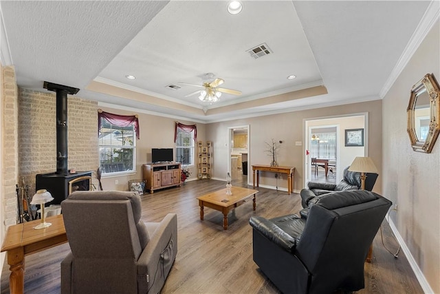 living room with a raised ceiling, ceiling fan, crown molding, hardwood / wood-style floors, and a wood stove