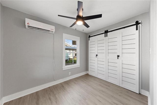 unfurnished bedroom featuring a wall mounted AC, ceiling fan, a barn door, light hardwood / wood-style floors, and a closet