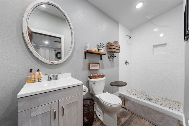 full bath with tiled shower, a textured wall, toilet, wood finished floors, and vanity