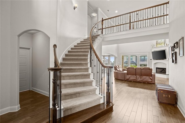 stairway with baseboards, arched walkways, a fireplace with raised hearth, a towering ceiling, and wood finished floors