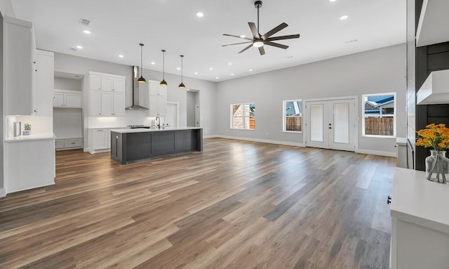 kitchen featuring ceiling fan, hanging light fixtures, wall chimney range hood, a kitchen island with sink, and white cabinets