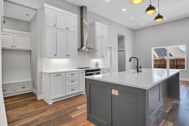 kitchen featuring a kitchen island with sink, sink, wall chimney range hood, white cabinets, and hanging light fixtures