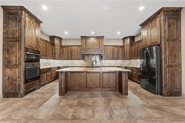 kitchen with backsplash, sink, stainless steel fridge, an island with sink, and light stone counters