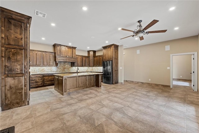 kitchen featuring stainless steel fridge, tasteful backsplash, custom exhaust hood, a kitchen island with sink, and ceiling fan