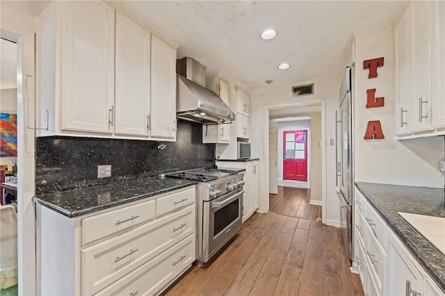 kitchen featuring dark wood-type flooring, stainless steel appliances, wall chimney range hood, tasteful backsplash, and dark stone counters