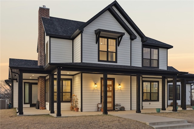 back of house at dusk featuring central AC unit, a chimney, a patio area, and a shingled roof