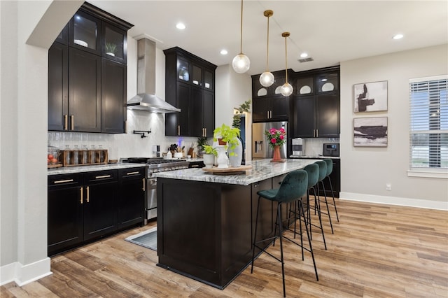 kitchen featuring a kitchen island with sink, stainless steel appliances, hanging light fixtures, wall chimney range hood, and glass insert cabinets