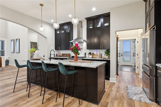 kitchen featuring a center island with sink, wall chimney exhaust hood, high quality fridge, glass insert cabinets, and light stone counters