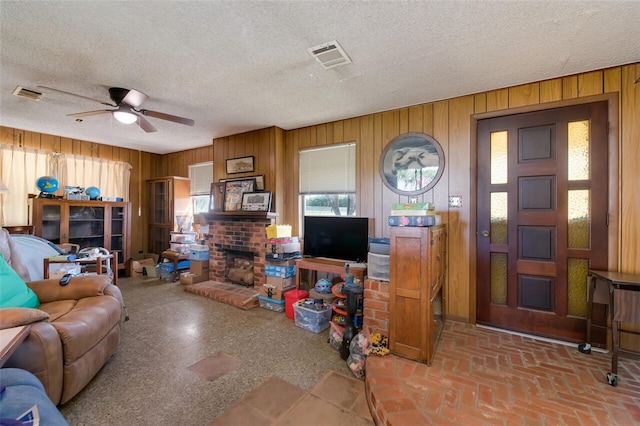 living room with wooden walls, ceiling fan, a textured ceiling, and a brick fireplace