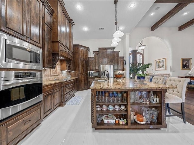 kitchen featuring appliances with stainless steel finishes, light stone counters, dark brown cabinetry, beamed ceiling, and an island with sink