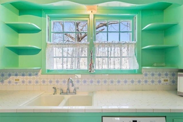 kitchen featuring open shelves, tile countertops, dishwasher, decorative backsplash, and a sink