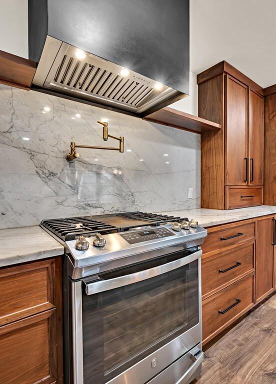 kitchen with stainless steel gas range oven, brown cabinetry, custom exhaust hood, and open shelves