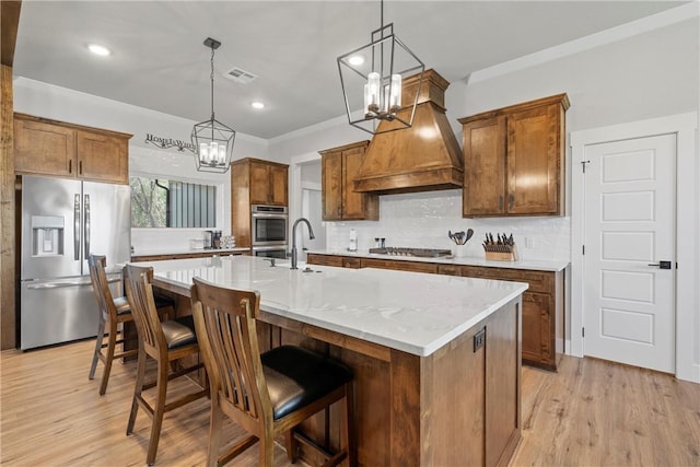 kitchen featuring light wood-type flooring, custom range hood, stainless steel appliances, a center island with sink, and hanging light fixtures