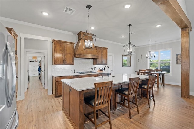 kitchen featuring appliances with stainless steel finishes, custom range hood, a kitchen island with sink, decorative light fixtures, and light hardwood / wood-style floors